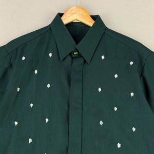 May include: Dark green button-down shirt with a classic collar and short sleeves. The shirt features a pattern of small, white diamond-shaped designs scattered across the fabric. The shirt is hanging on a wooden hanger.