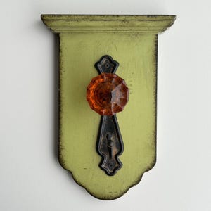May include: A wall-mounted hook featuring a light green wooden plaque with distressed dark edges. A black metal backplate is centered on the wood, holding a faceted, translucent orange glass knob at the top and a small metal hook at the bottom.