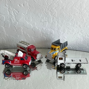 May include: Four toy trucks in red, yellow, and silver. One red truck has a tow truck, another has a tank, and a third has a dump bed. The fourth truck is silver with a flatbed. The red truck has the text "24 HR SERVICE".