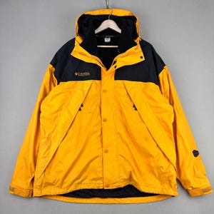 May include: A yellow and black Columbia hooded jacket. The jacket has a black hood and shoulders, with a yellow body. It features a zipper and button closure, and the Columbia logo is visible on the chest. The jacket is hanging on a hanger.