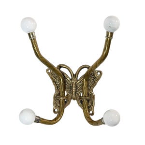 May include: A decorative wall hook with a butterfly design. The hook is made of brass-colored metal and features a butterfly-shaped backplate. Four curved arms extend from the backplate, each ending in a white, spherical knob. The arms are textured with a vine-like pattern.