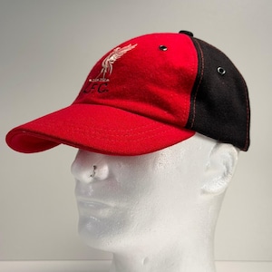 May include: Red and black baseball cap with the Liverpool Football Club logo embroidered on the front. The cap features a red brim and front panel, with a black back panel. The logo is a white bird with the letters "LFC" below it.
