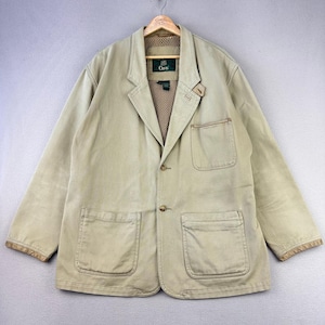 May include: A tan blazer with two large front pockets and a notched lapel. The jacket has a button closure and a brown mesh lining. The brand name "Orvis" is visible on the label. The jacket is hanging on a wooden hanger.