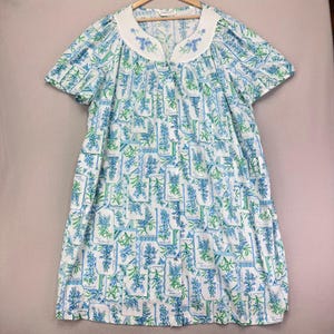 May include: A short-sleeved, white and blue floral print dress. The dress features a white collar with blue floral embroidery. The fabric has a repeating pattern of blue flowers and green leaves on a white background. The dress is hanging on a wooden hanger.