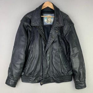 May include: Black leather jacket with a collared neckline, zipper closure, and multiple pockets. The jacket has a vintage look and a label with a portrait. The jacket is hanging on a wooden hanger.
