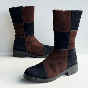 May include: A pair of mid-calf boots featuring a patchwork design of dark brown and black suede panels. The boots have a rounded toe, a flat black rubber sole with a low block heel, and a vertical seam construction that alternates the two colors.