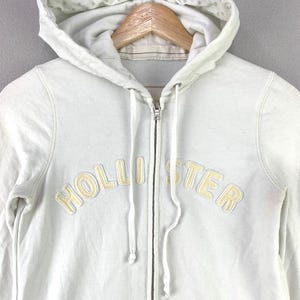 May include: A white zip-up hoodie featuring the word HOLLISTER in light yellow embroidered lettering across the chest. The garment includes a drawstring hood with a white interior lining and a full-length metal zipper closure down the center.