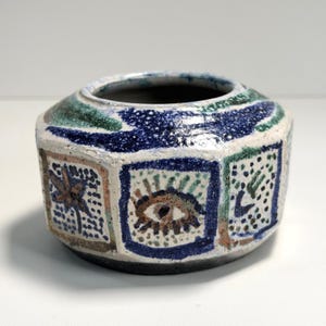May include: A handcrafted ceramic vase with a unique, geometric shape. The vase features a textured, off-white base with blue, green, and brown painted designs. The exterior is decorated with square panels, each containing a different abstract motif.