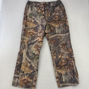 May include: Camouflage pants in shades of brown, green, and tan, featuring a leaf and branch pattern. The pants have a button and zipper closure, pockets, and a relaxed fit. Ideal for outdoor activities or hunting.