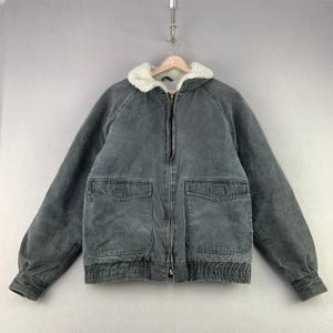 May include: A charcoal gray zip-up jacket featuring a white sherpa-lined collar, long sleeves with ribbed cuffs, and two large front patch pockets with flaps. The jacket has a full-length front zipper closure and a ribbed hem at the waist.