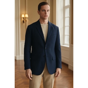 May include: A navy blue blazer with a two-button closure, paired with tan pants and a light beige crew neck shirt. The blazer has a classic lapel and a tailored fit. The outfit is completed with a pair of tan pants.