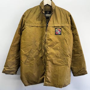 May include: A tan-colored jacket with a full-length zipper and a small patch on the upper right chest. The jacket has a collared neckline and two front pockets. The patch features a crest design with red, white, and black elements.