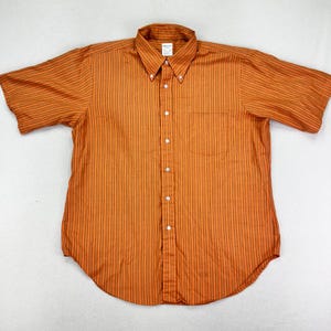 May include: Orange short-sleeve button-down shirt with vertical orange and red stripes. The shirt has a button-down collar, a chest pocket, and white buttons. The shirt is laid flat on a white surface.