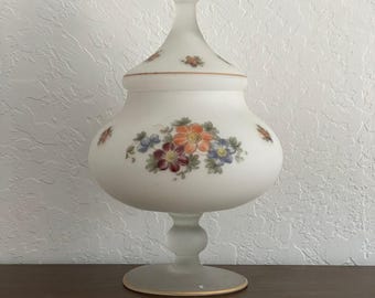 May include: A frosted glass apothecary jar with a lid, featuring a floral design in shades of orange, red, blue, and yellow. The jar has a bulbous body and a pedestal base, with a gold rim on the base and lid.