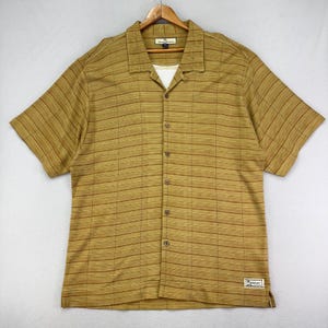 May include: A short-sleeved button-down shirt in a golden-brown color with a striped pattern. The shirt has a collared neckline and a white undershirt is visible. A small label is sewn on the bottom right side of the shirt.