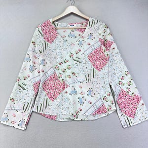 May include: A long-sleeved blouse with a V-neck and a patchwork design. The blouse features a mix of floral patterns in pink, blue, and white. The sleeves are long and flared, and the blouse is hanging on a wooden hanger.