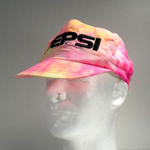 May include: A tie-dye visor in shades of pink, yellow, and orange. The word "PEPSI" is printed across the front in black block letters. The visor is made of a lightweight material and has a curved brim.