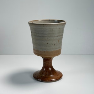 May include: A handmade ceramic goblet with a wide, slightly flared rim. The upper portion is a muted gray with subtle speckles, transitioning to a warm brown base and stem. The goblet stands on a round, stable base.
