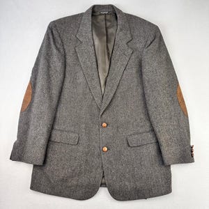 May include: A gray tweed blazer with brown leather elbow patches. The blazer has two front pockets, a notched lapel, and two brown buttons. The sleeves have three brown buttons. The interior lining is a light brown color.