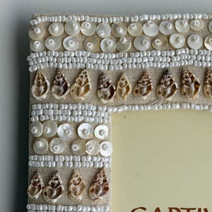 May include: Close-up of a decorative photo frame with a beige linen fabric. The frame is embellished with rows of white beads, iridescent shell-shaped accents, and round mother-of-pearl sequins. The frame has a neutral color palette.