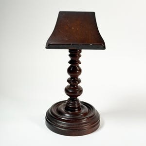 May include: A dark brown wooden stand with a square top and a turned, baluster-shaped stem. The base is round and has a decorative, layered design. The stand is likely for displaying an object.