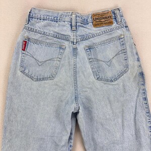 May include: Light blue denim jeans with a high waist and classic five-pocket design. The back features two pockets with decorative stitching and a brown leather patch with the text "UNIONBAY". A red tag is visible on the back pocket.