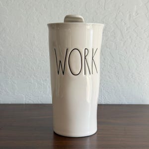May include: A white ceramic container with a lid, featuring the word "WORK" in black lettering. The container has a smooth, glossy finish and is set against a neutral background.
