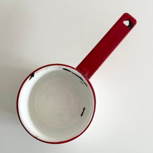 May include: A small, red saucepan with a long handle. The interior is white, and the rim is red. The handle has a hole at the end for hanging. The pan shows signs of wear, with some black marks on the rim.