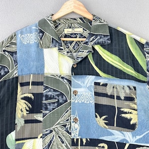 May include: A patchwork button-down shirt with a tropical print. The shirt features a variety of patterns, including palm trees, leaves, and geometric shapes in shades of blue, green, and black. The shirt has a collared neckline and a single pocket.
