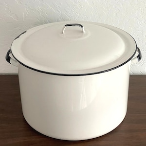 May include: A large, white enamel pot with a matching lid. The pot has a black rim and handles, and a black handle on the lid. The pot is sitting on a wooden surface. A classic kitchenware item.