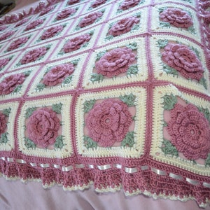 May include: A handmade crochet blanket featuring a repeating pattern of pink roses on a cream and pink background. The blanket has a ruffled edge with white ribbon accents. The overall design is a floral theme.