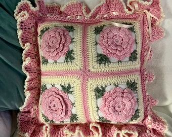 May include: A crocheted pillow with a pink and cream floral design. The pillow has a ruffled edge and a satin ribbon.
