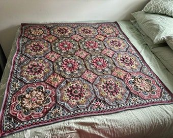 May include: A crocheted throw blanket with a geometric pattern in shades of pink, brown, and gray. The blanket is made up of repeating hexagonal motifs.