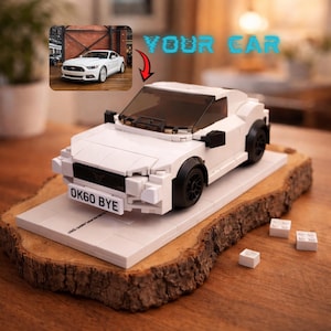 Op de afbeelding: Een witte speelgoedauto gemaakt van bouwstenen, met zwarte wielen en een donkere voorruit. De auto staat op een witte basis op een houten plank. De nummerplaat luidt "OK60 BYE". De woorden "YOUR CAR" staan boven de auto.