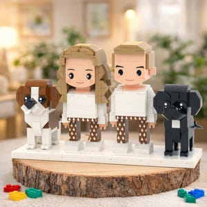 Puede incluir: Un retrato familiar estilo Lego con dos figuras y dos figuras de perro. Las figuras tienen cabello rubio y castaño claro, tops blancos y pantalones estampados. Los perros son marrones y blancos, y negros. Se paran en una base blanca sobre una rodaja de madera.