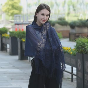 May include: A navy blue shawl with intricate floral lace detailing along the edges and a fringed hem. The shawl is draped over the shoulders, creating a flowing, elegant look. The fabric appears lightweight and semi-sheer, suitable for layering.