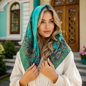 May include: A square headscarf featuring a vibrant teal background with a detailed paisley pattern in shades of gold, black, and white. The fabric is draped over the head, showcasing intricate floral and teardrop motifs across the entire surface.