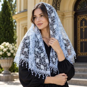 May include: A light blue lace chapel veil featuring a delicate floral pattern and scalloped edges with small dangling bead accents. The triangular head covering is draped over the head and shoulders, showcasing intricate embroidery and sheer fabric.
