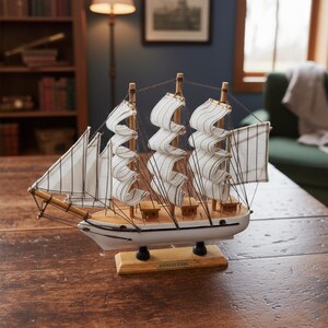 May include: A detailed model sailboat with white sails and a wooden hull. The sails are striped, and the boat is mounted on a wooden base. The model is displayed on a dark wooden table, showcasing nautical decor.