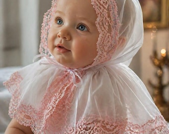 Può includere: Un bambino con un cappuccio e un colletto bianchi rifiniti con pizzo rosa. Il bambino ha gli occhi azzurri e guarda in alto. Il cappuccio e il colletto sono realizzati in un tessuto trasparente. L'estetica generale è morbida e delicata.