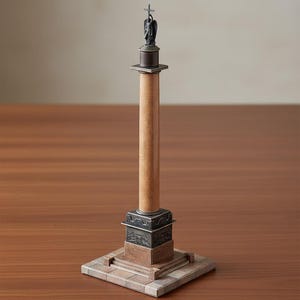 May include: A detailed miniature column replica. The light brown pillar has a dark bronze top and base, with a square, detailed base. A small statue of an angel holding a cross is at the top. The column is set on a square base, reminiscent of a monument.