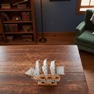 May include: A detailed wooden model of a sailing ship with three masts and white sails. The ship is painted white and brown, with blue accents. The model sits on a wooden base, and is displayed on a dark wooden table.