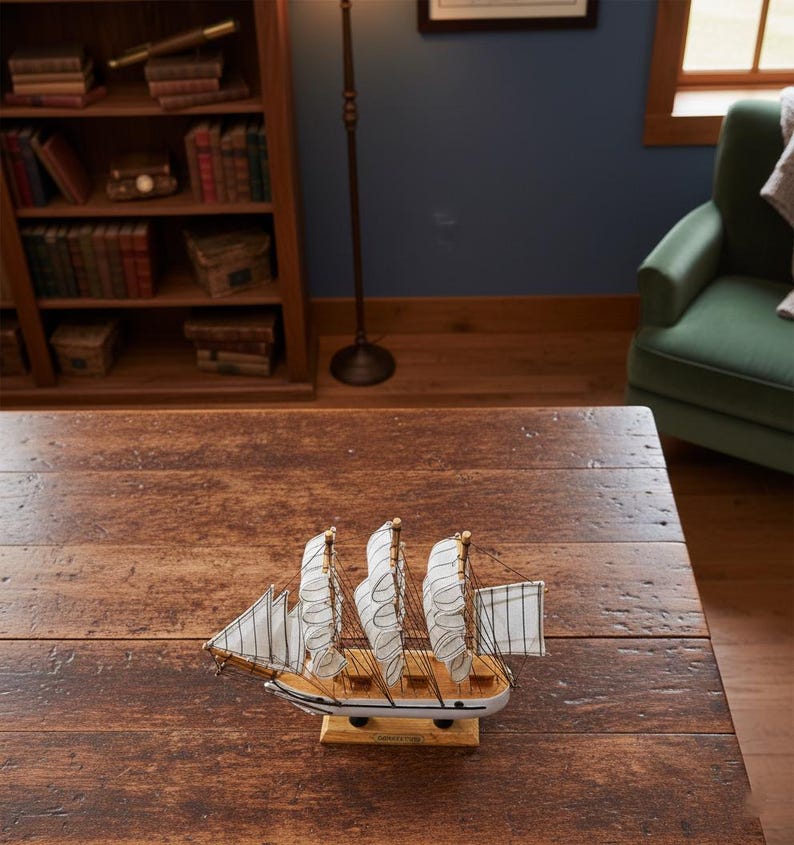 May include: A detailed wooden model of a sailing ship with three masts and white sails. The ship is painted white and brown, with blue accents. The model sits on a wooden base, and is displayed on a dark wooden table.