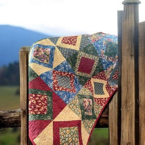 May include: A colorful patchwork quilt draped over a wooden fence. The quilt features squares of floral, geometric, and solid fabrics in shades of red, green, blue, and cream. The quilt's design is a mix of patterns and textures.