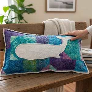May include: A rectangular decorative pillow with a patchwork design in shades of blue, teal, and purple. A white whale applique is centered on the pillow. The pillow is placed on a wooden bench.