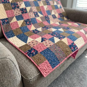 May include: A patchwork quilt featuring a repeating geometric pattern of squares and triangles. The fabric displays a mix of floral prints and solid colors in shades of dusty pink, navy blue, and tan, finished with a pink fabric binding edge.