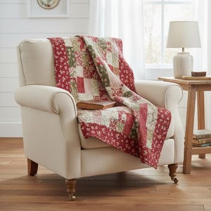 Könnte beinhalten: Ein cremefarbener Sessel mit einer rot, grün und weiß gefleckten Patchwork-Decke. Ein Buch liegt auf der Decke. Eine weiße Lampe und ein kleiner Holztisch befinden sich im Hintergrund und schaffen eine gemütliche, einladende Szene.