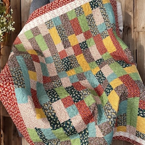 Op de afbeelding: Een kleurrijke patchworkquilt met rechthoekige blokken in rood, groen, geel en blauw. De quilt heeft bloempatronen en een rode rand met een wit bloemmotief. De quilt is over een stoel gedrapeerd.