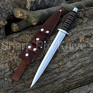 May include: A dagger with a double-edged, silver blade and a handle wrapped in alternating black and brown bands. It rests beside a brown leather sheath with white rivets. The dagger is displayed on a textured wooden surface.