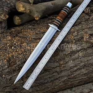 May include: A dagger with a polished steel blade and a handle featuring alternating bands of brown wood and black material. A metal ruler, marked in inches, is positioned alongside the blade. The dagger's total length is approximately 12 inches.